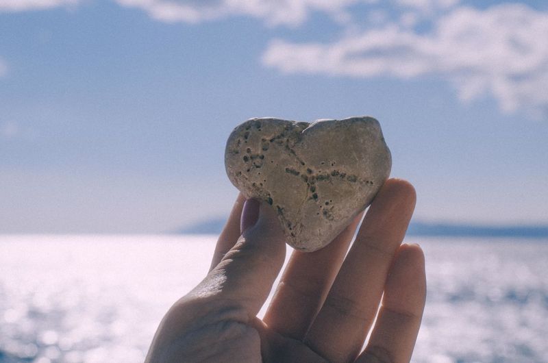 Close-up of hand holding heart shape rock against sky
