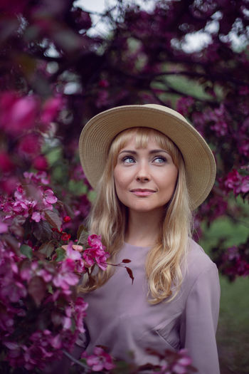 Portrait young beautiful woman in a pink dress and hat stands by a blooming pink apple tree