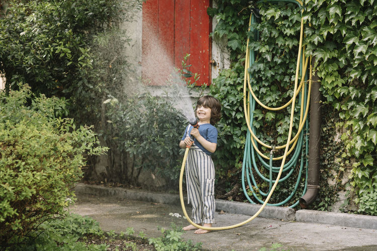 Boy enjoying holding garden hose while watering plants at backyard