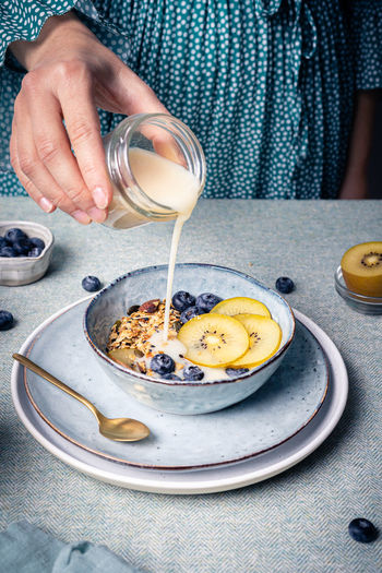 High angle of crop anonymous female adding milk into bowl with healthy muesli with blueberries and pieces of kiwi for healthy breakfast