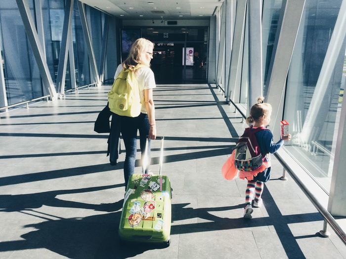 Rear view of mother pulling suitcase while walking with daughter at airport corridor