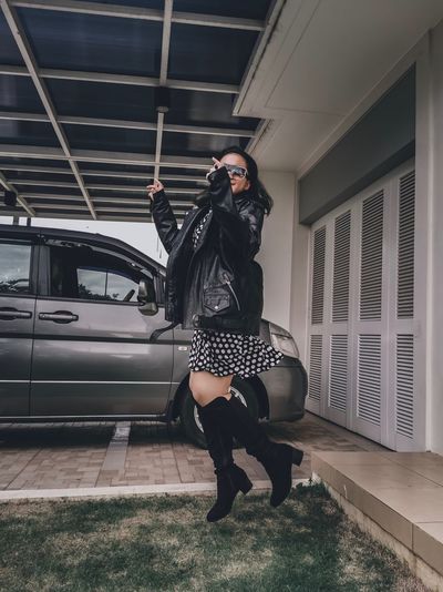 A girl wearing a dress, jacket, sun glasses and boots jumping off the porch to the grass 