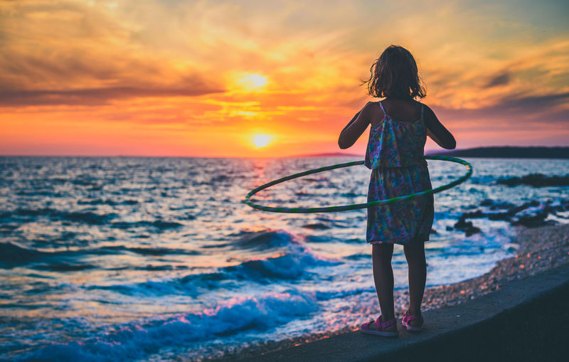 Full length of girl playing with hula hoop on shore at beach against sky during sunset