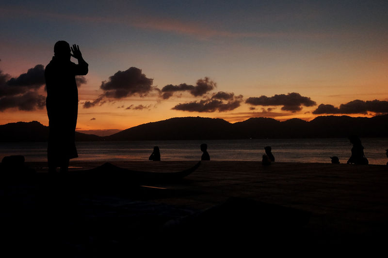 Silhouette people praying by sea against sky during sunset