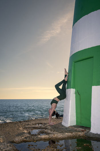 Sporty woman doing a handstand against the wall of a lighthouse by the ocean.