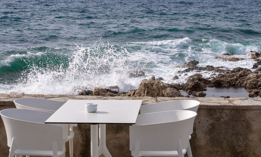 Empty table and chairs against waves splashing on rocks in sea