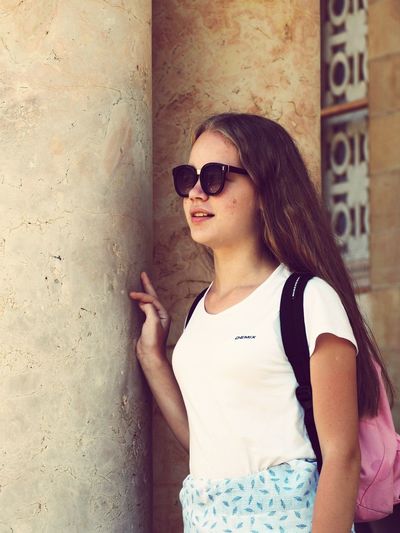Girl with backpack wearing sunglasses while standing against wall
