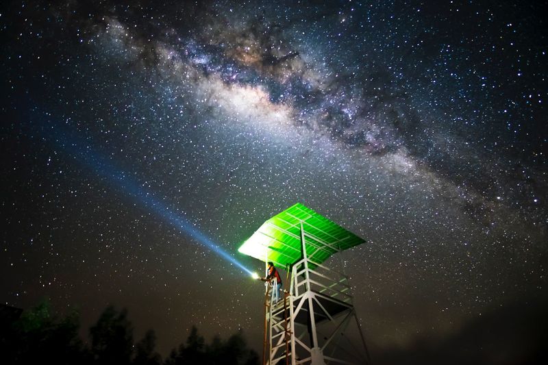 Low angle view of man holding flashlight while standing at observation point against sky during night