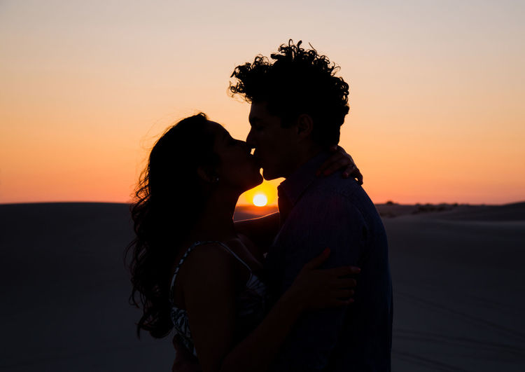 Side view of couple kissing on desert against sky during sunset
