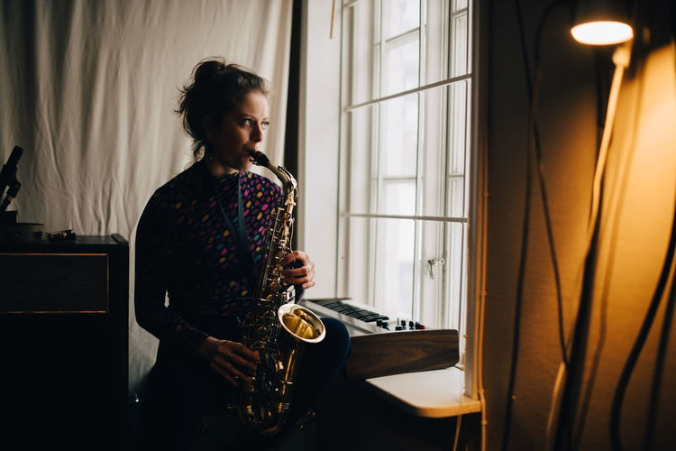 Young woman playing saxophone while practicing by window at studio