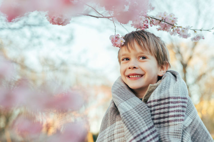 Portrait of a little boy wrapped in a blanket enjoying cherry blossoms in a city park.