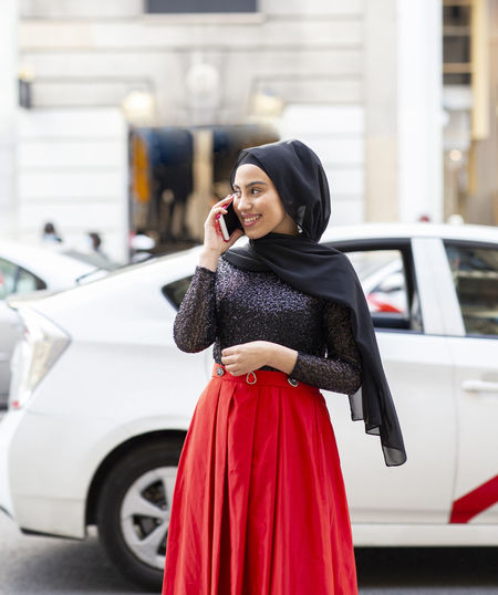 Portrait of young beautiful woman wearing black hijab talking on smart phone in middle of street