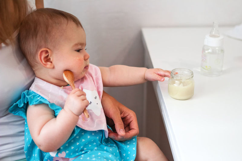 Baby girl holds wooden spoon in hand and tries to eat herself vegetable puree from glass jar.
