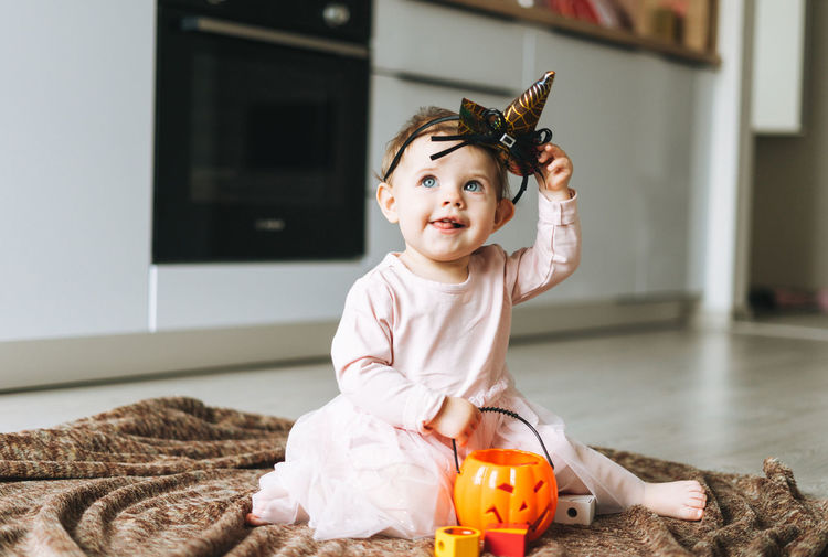 Cute little baby girl in witch's hat and pumpkin lantern in kitchen at home, halloween time