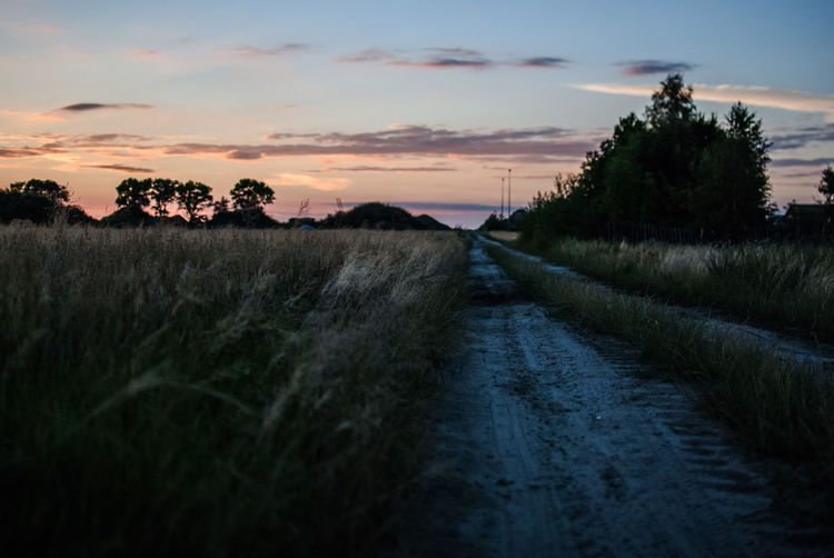 Dirt road amidst field against sky during sunset