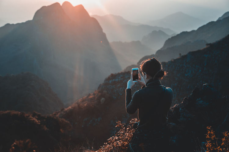 Rear view of woman photographing mountains through mobile phone during sunset