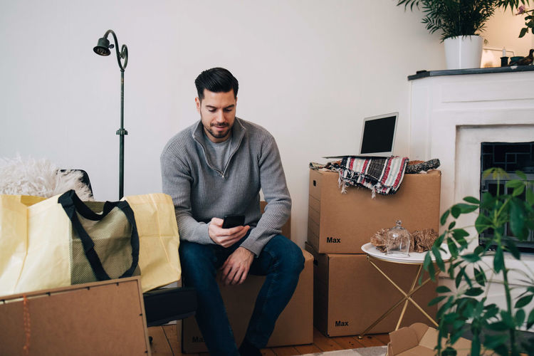 Mid adult man using mobile phone while sitting on box at home