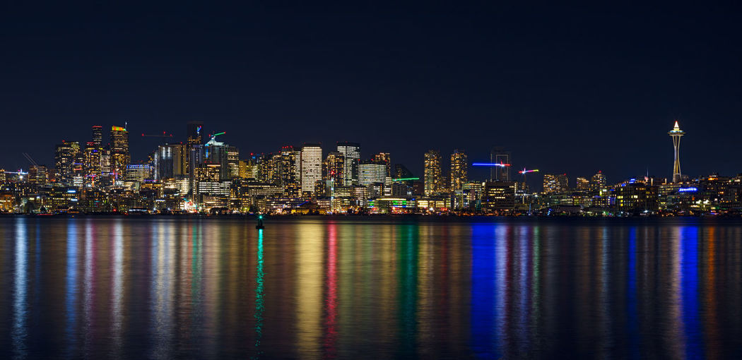 Illuminated buildings by sea against sky at night