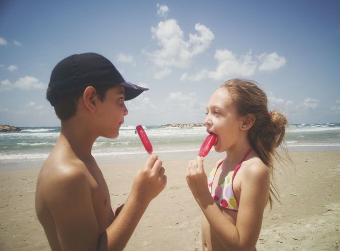 Side view of siblings eating popsicles while standing face to face at beach