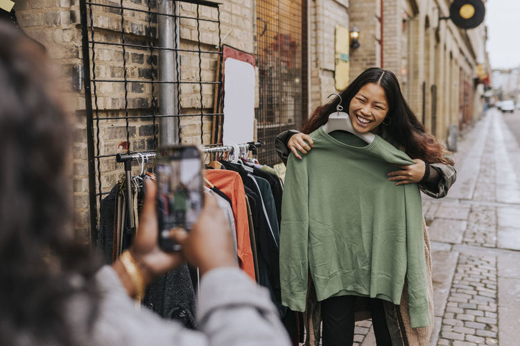 Mature woman photographing cheerful female friend trying on clothes at street