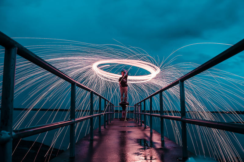 Man spinning wire wool on footbridge against sky at night