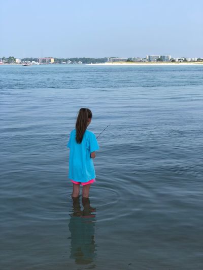 Rear view of girl fishing while standing in sea against sky