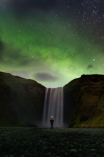 Back view of unrecognizable tourist admiring amazing scenery of powerful skogafoss waterfall flowing through rocky cliff under magnificent starry sky with polar lights during trip in iceland person