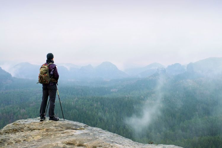 Hiker with backpack and poles on cliff rock watching over the misty and foggy spring valley.