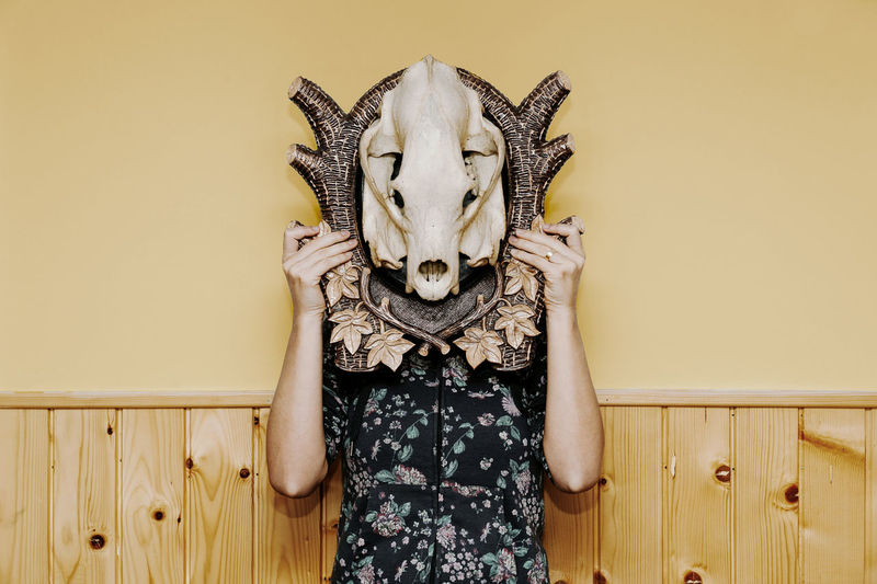 Woman with face covered by animal skull standing against wall
