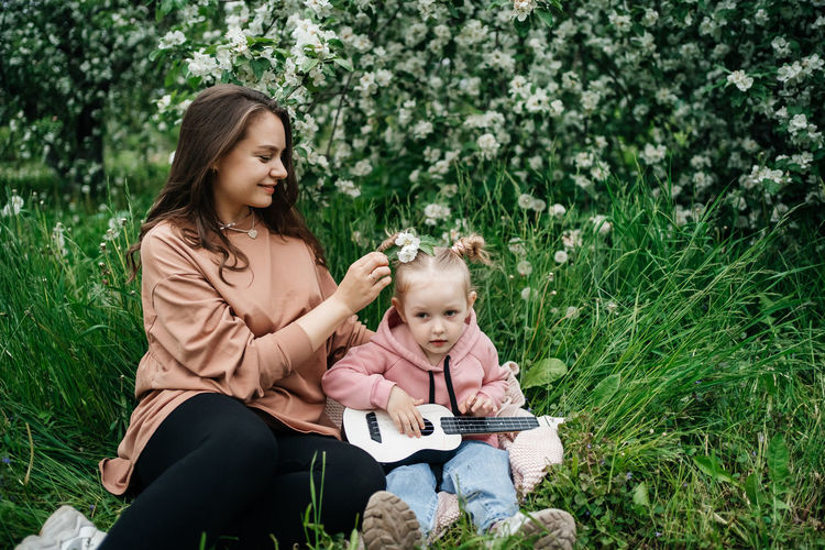 Young mother and baby daughter play the ukulele in a blooming apple orchard in nature