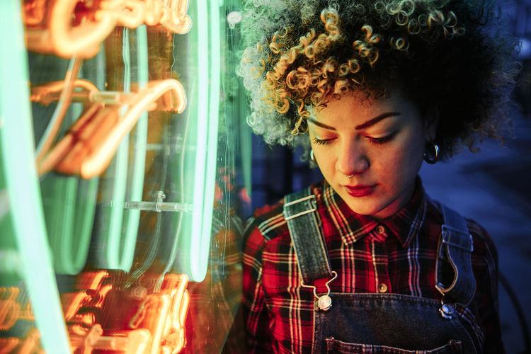 Curly haired woman looking down while standing by neon lights