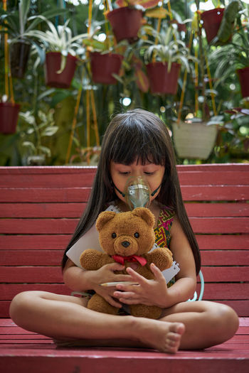 Girl wearing oxygen mask while sitting with teddy bear on bench