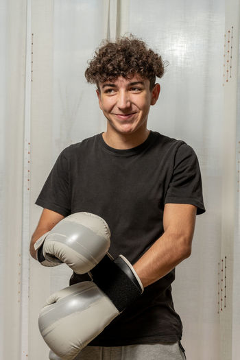 Smiling young man wearing boxing glove standing against wall