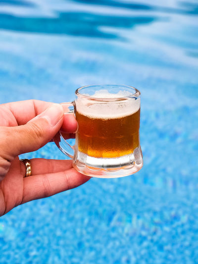 Hand holding a shot glass size mug of beer with pool in background