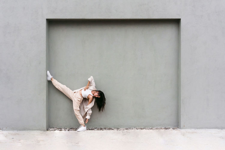 Creative cool female leaning on gray wall and dancing expressively in city street
