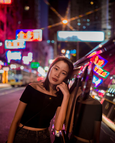 Portrait of young woman standing by car against illuminated building