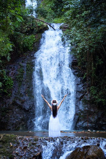 Rear view of young woman with arms raised standing by waterfall