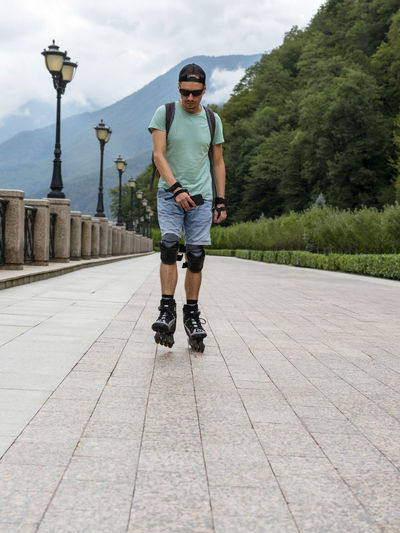 Young man in protective equipment roller skating along embankment against forest mountains in summer