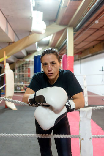 Real female boxer in gloves leaning on rope at ring before training