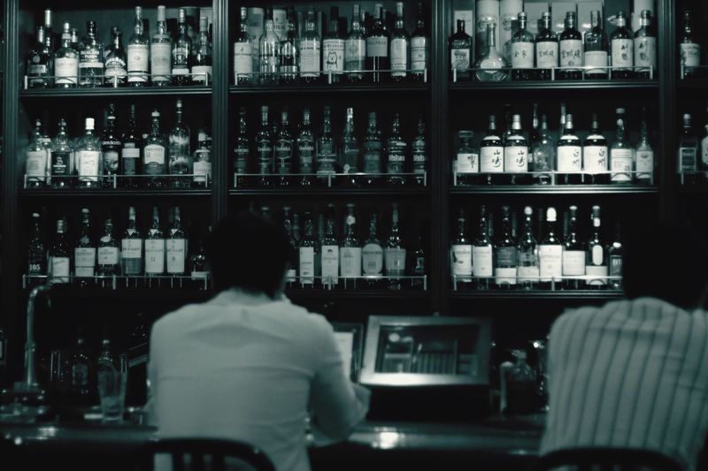 Rear view of men sitting against alcohol bottles on rack in bar