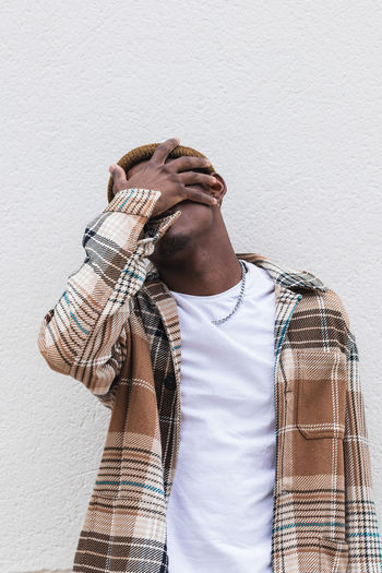Unrecognizable young black man in checkered shirt covering face with hand while standing against gray wall on city street