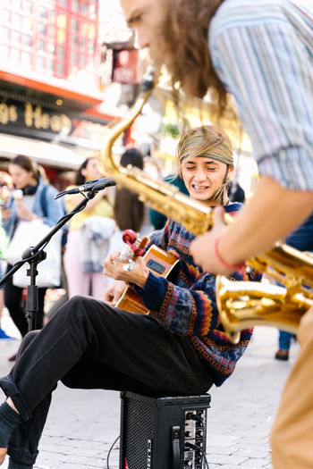 Selective focus ont he face of a man singing while playing guitar next to a saxophonist in the street