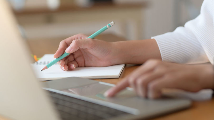 Cropped hands of woman using laptop while writing in diary