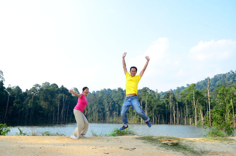 Cheerful people with arms outstretched jumping by lake against forest and sky