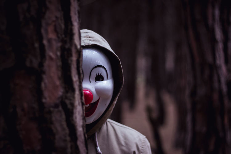 Person wearing mask and hood by tree trunk in forest