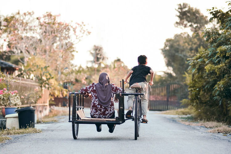 Rear view of boy riding bicycle with mother on road
