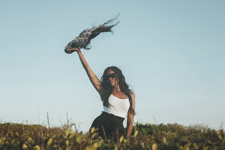 Low angle view of woman with arm raised holding scarf against sky