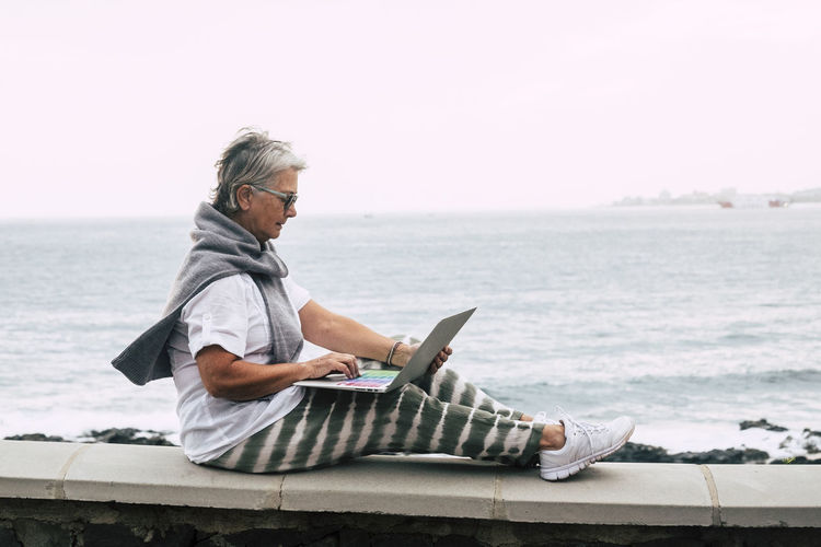 Side view of man using laptop while sitting on retaining wall against sea