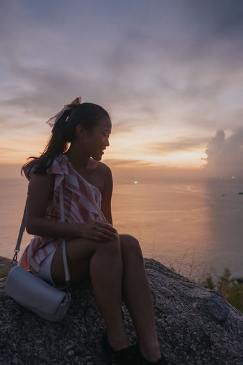 Side view of young woman sitting on beach against sky during sunset