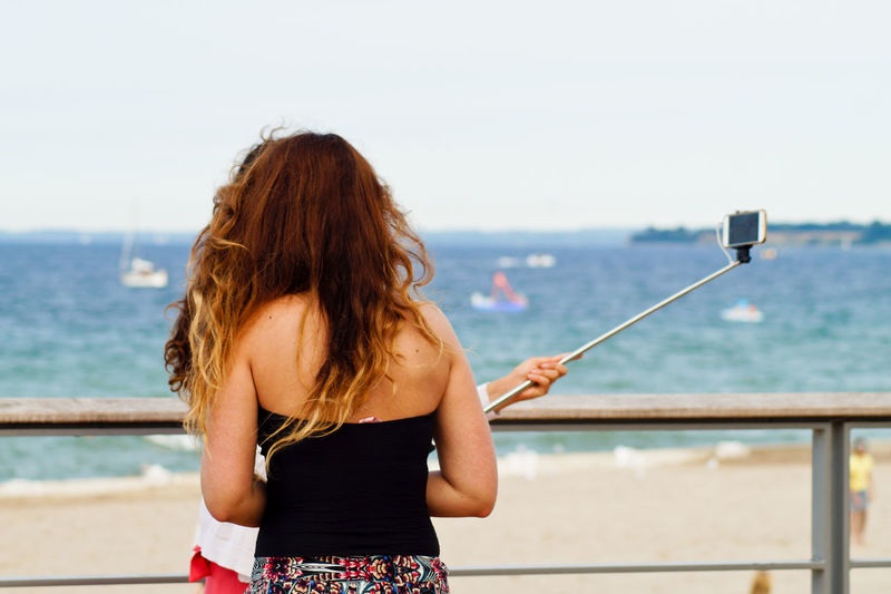 Rear view of friends taking selfie while standing by railing against sea